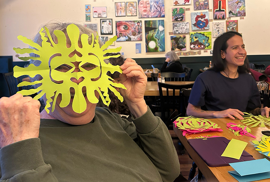 Smiling senior in art-filled cafe holding a yellow hand-cut paper mask in front of their face.  Photo credit Witt Siasoco.
