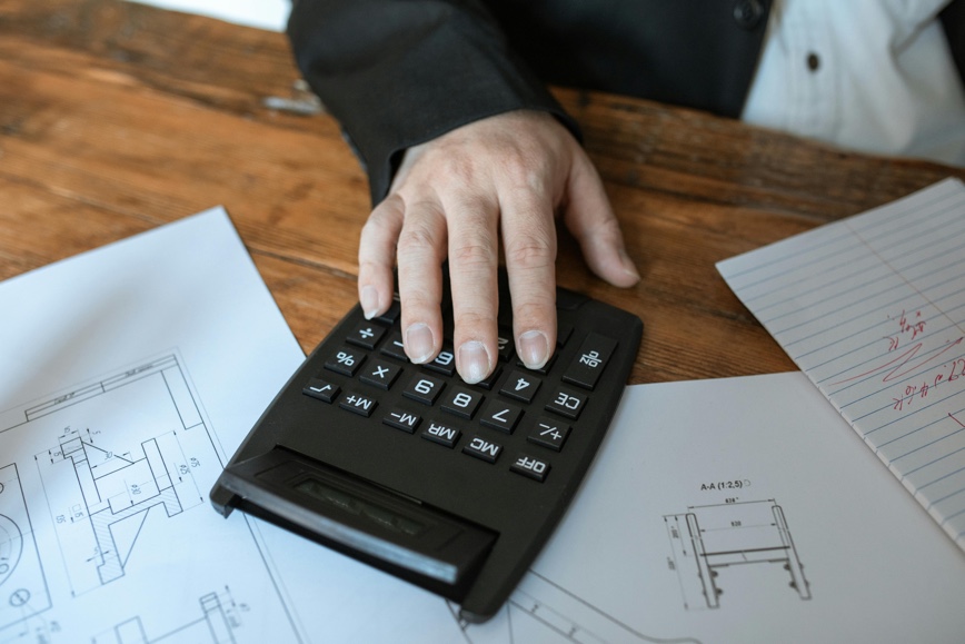 Desk with blueprints, calculator and a person's hand on top.