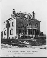 Black and white exterior photo of two sotry brick mansion with large fron porch and balcony.