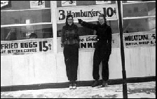 Black and white photo of two people standing outside in front of diner window  by sign that reads three hamburgers for 10 cents. 1939