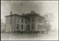 Faded sepia exterior image of the two-story school building with students gathered on the lawn in the foreground.