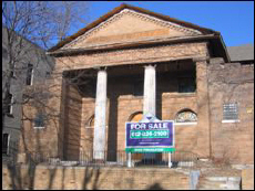 Color photo of the front of the church showing red brick masonry, columns, gabled roof and a for sale sign in the forefront.